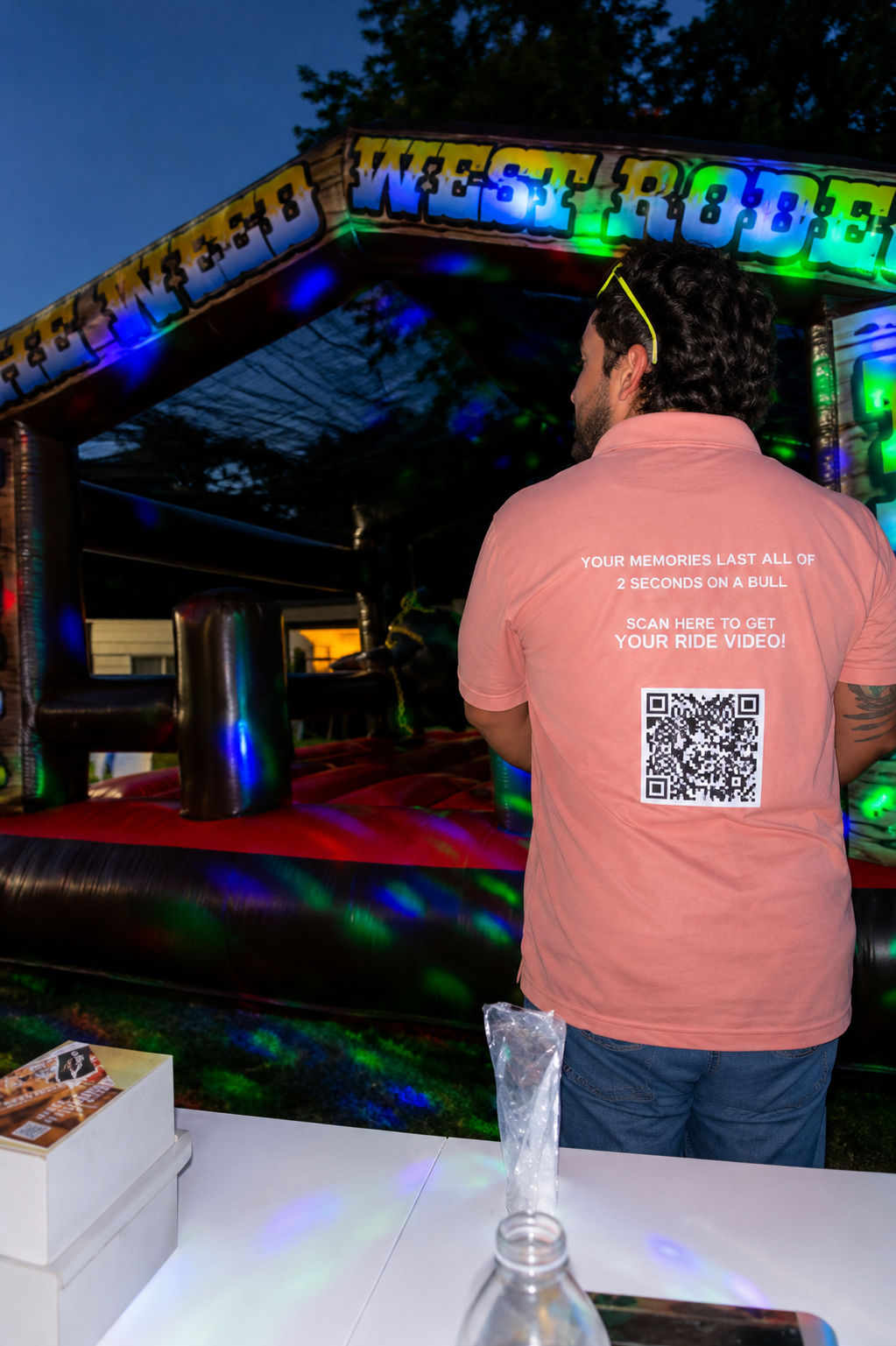 Staff member in a Wild West Rodeo polo standing in front of the glowing mechanical bull at dusk
